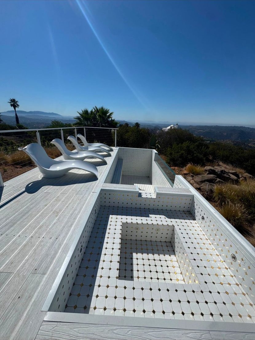 A white tiled plunge pool and spa on a deck with lounge chairs, hills in the distance.